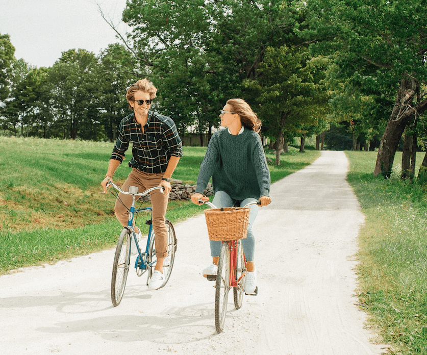 A man and woman enjoy an effortless, fun bike ride in classic, high-end casual wear, embodying a cool, confident lifestyle.