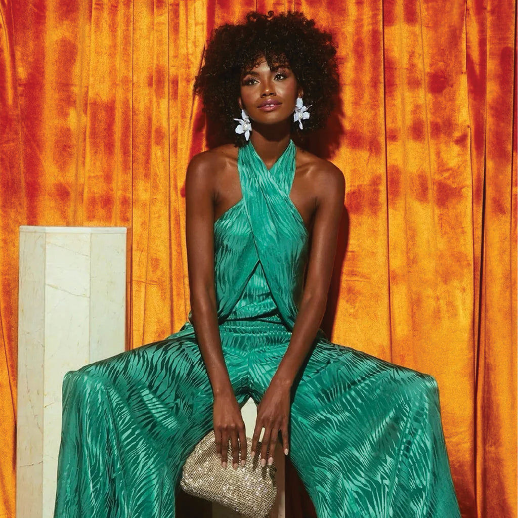 A classic, dark-skinned model in an effortless, bright green halter top and trousers, holds a sparkly gold clutch, embodying high-end, fun fashion.