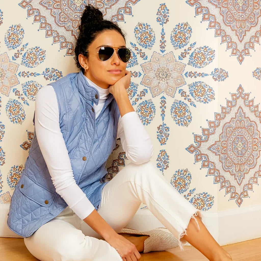 Woman in aviator sunglasses, light blue quilted vest, and frayed-hem jeans, embodying an effortless, high-end style against a mandala-patterned wallpaper.