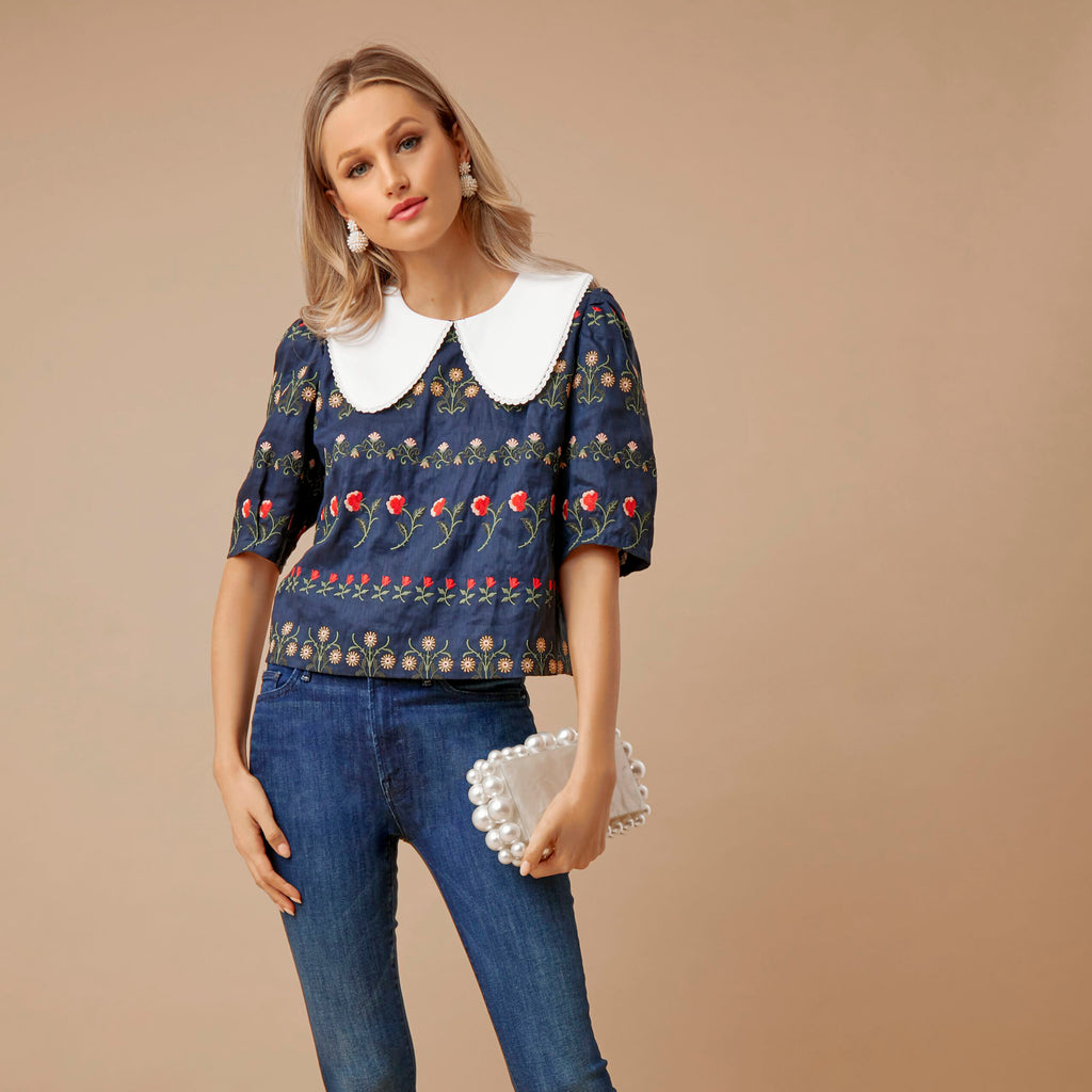Model in slim-fit jeans and a navy blouse with floral embroidery, classic Peter-Pan collar, holding a pearl-adorned clutch; embodies effortless, high-end style.