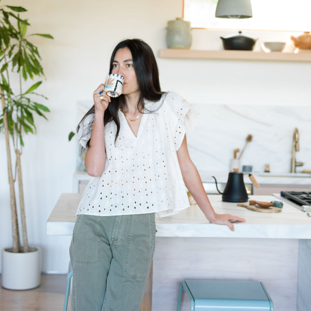 Woman in a classic white eyelet blouse and olive-green pants, sipping from a patterned mug, with high-end kitchen accessories like a gooseneck kettle nearby.