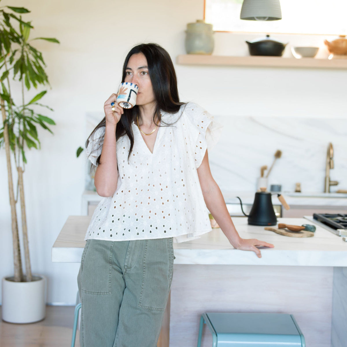 Woman in a classic white eyelet blouse and olive-green pants, sipping from a patterned mug, with high-end kitchen accessories like a gooseneck kettle nearby.