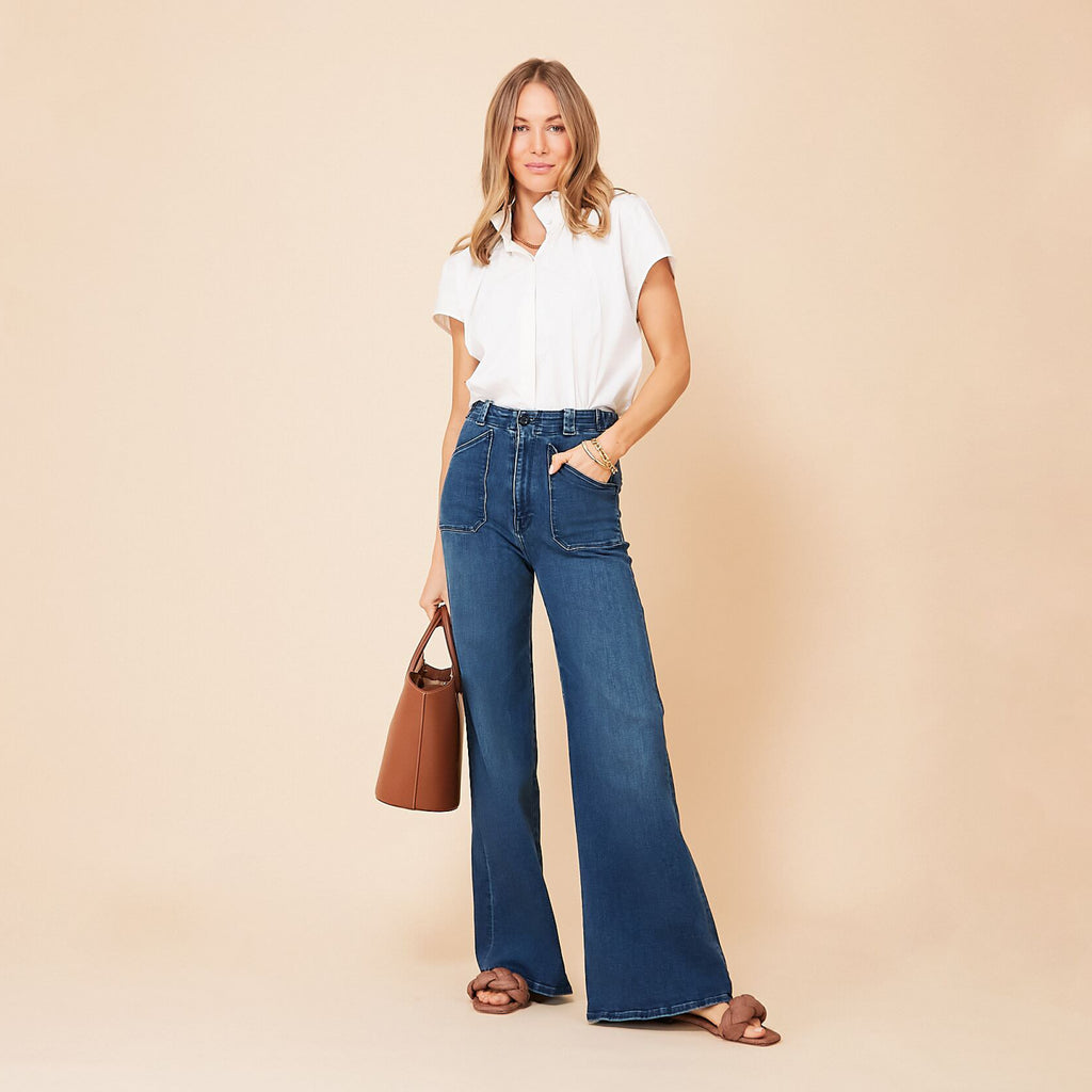Woman in a classic white button-down and high-waisted blue jeans, carrying an effortless brown leather tote, styled with fun gold bracelets and braided sandals.