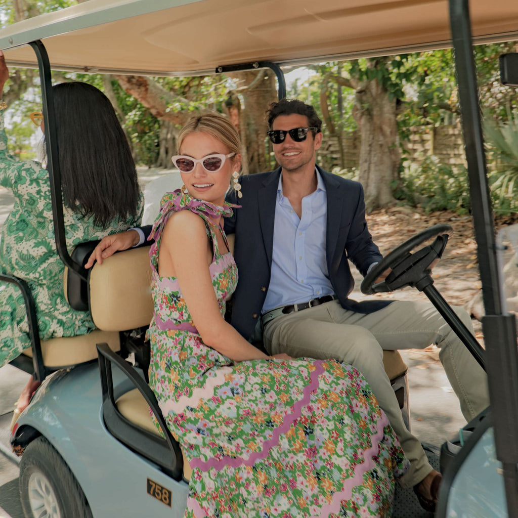 A classic light-blue golf cart carries a stylish couple; she wears a floral maxi dress, he in a navy blazer, embodying effortless, high-end fun.