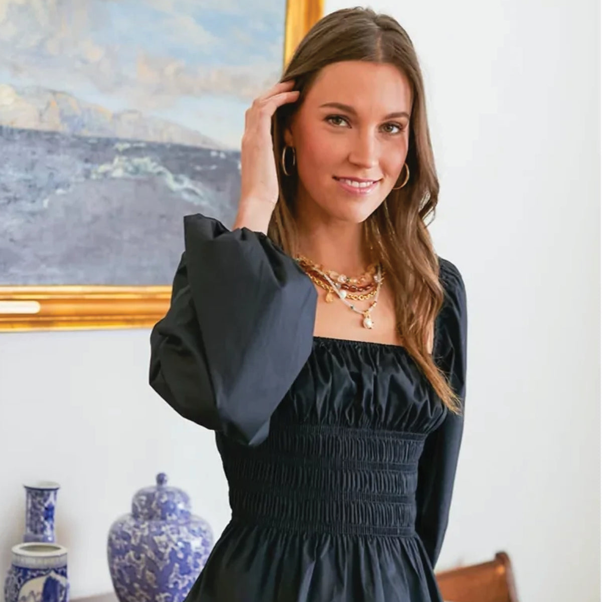 Young woman in a classic black smocked-waist dress with gold accessories, standing indoors before a seascape painting and decorative porcelain vases, exuding effortless elegance.