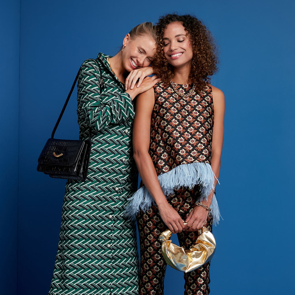 Two women in classic, high-end outfits; one in a green woven-pattern dress, the other in an effortless floral set. Both exude fun, confident elegance.