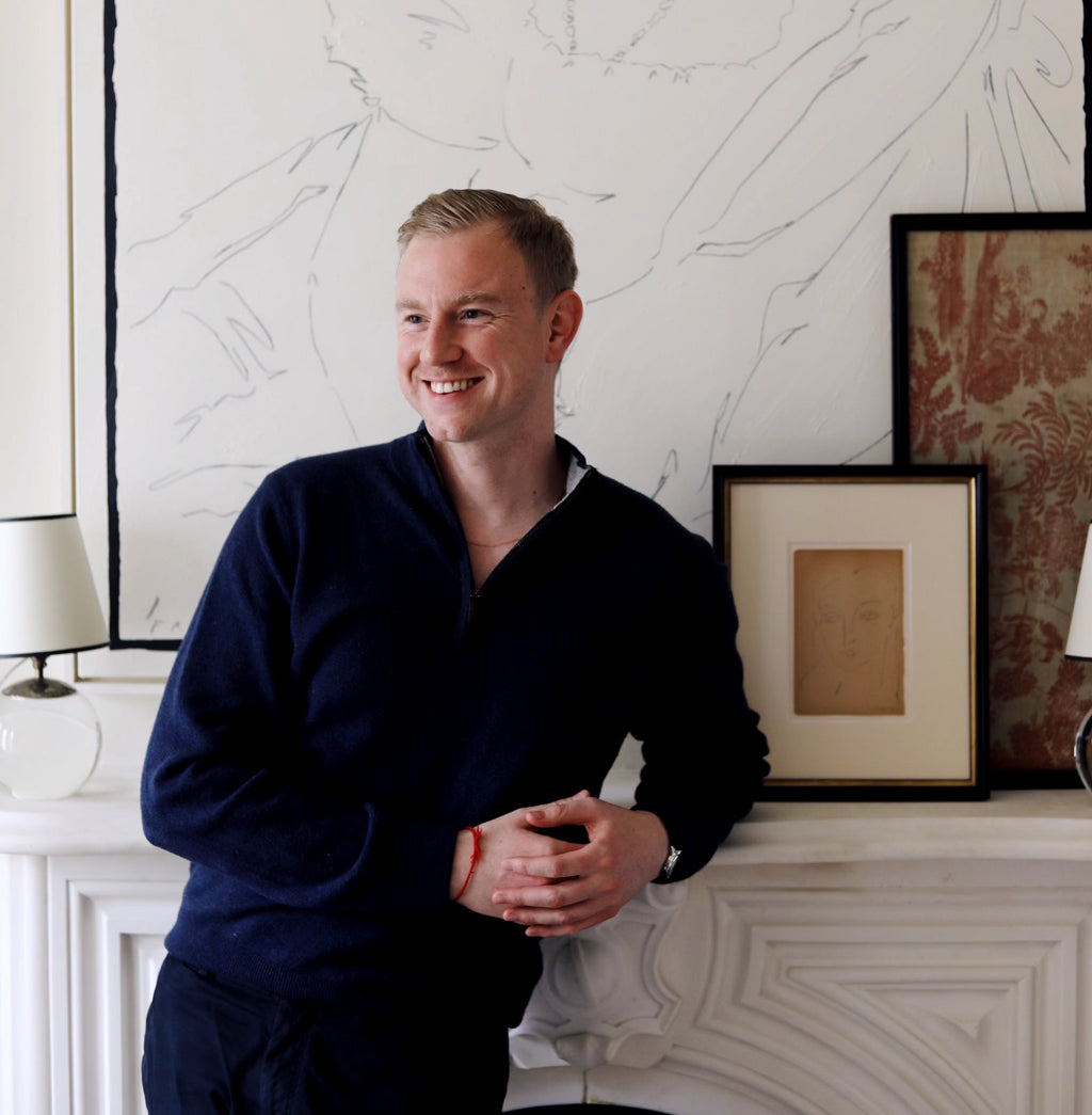 Young man in a classic living room, leaning on a mantel, wearing an effortless pullover and bracelet, surrounded by fun, high-end art and decor.