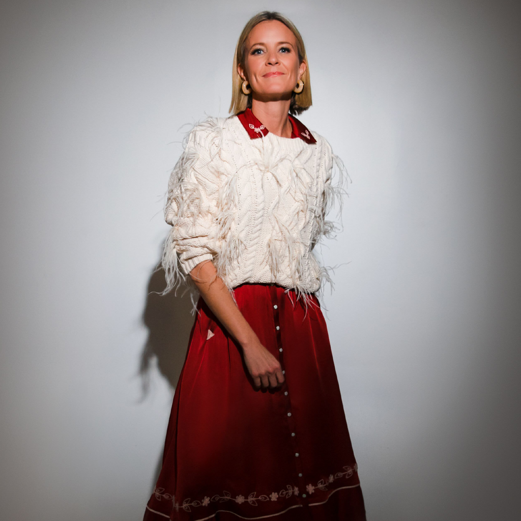 Woman in a classic, chunky white cable-knit sweater with fringe over a red floral-embroidered dress, embodying effortless, fun, and high-end style.