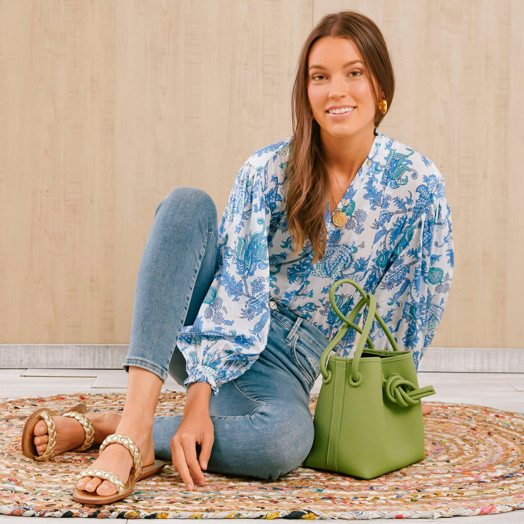 Young woman in classic blue jeans, floral blouse, and gold sandals sits on a braided rug with an effortless, high-end green tote beside her.