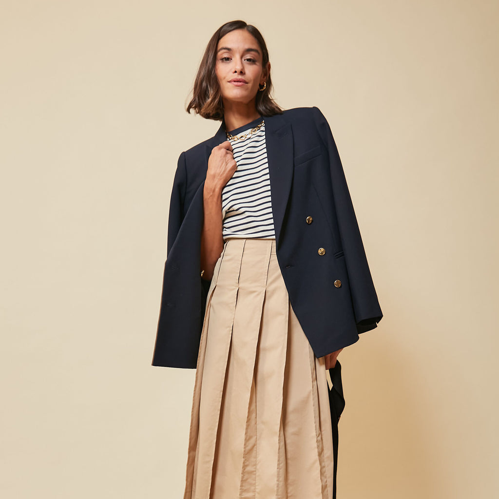 Young woman wearing a navy blazer, striped top, pleated tan maxi skirt, accessorized with gold jewelry; embodies classic, effortless, and high-end nautical style.