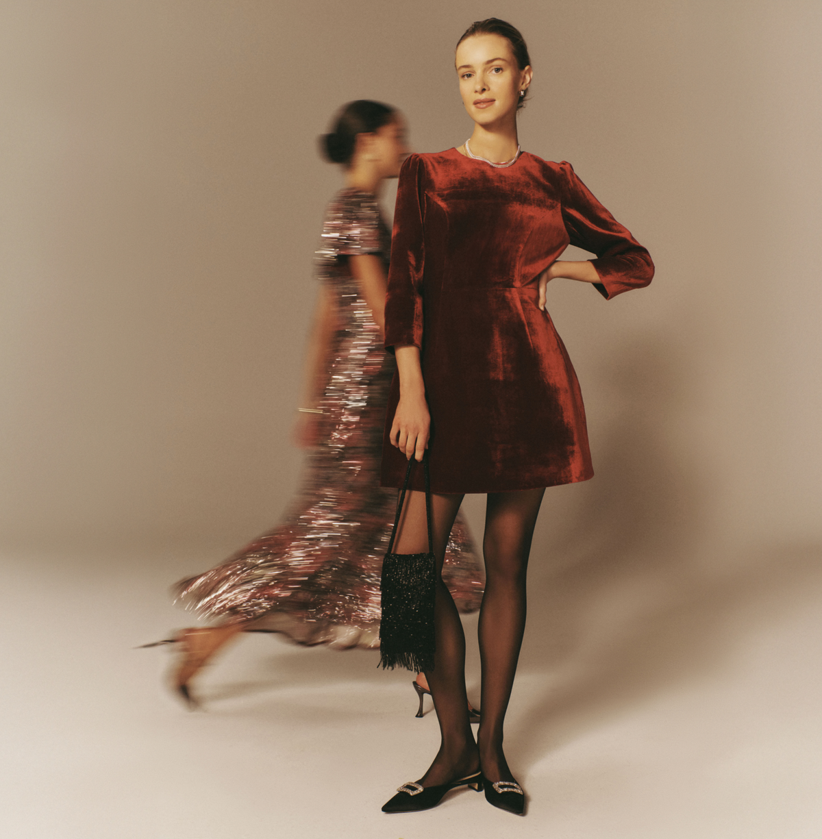 Woman standing in a red velvet dress with a woman walking in a sequined dress in the background.