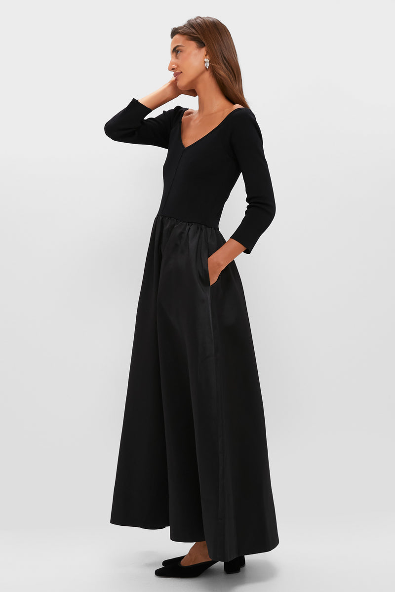 A woman in profile wears a Tuckernuck black fit-and-flare gown with long sleeves, a fitted top, flared skirt, and black heels, set against a plain white background.