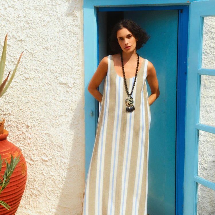Woman standing in front of a blue door in a long brown, blue, and white striped sleeveless tank dress with a long necklace.