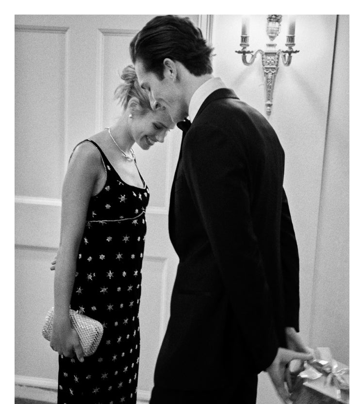A black and white image of a man and woman. The woman wears a sleeveless midi dress with crystal embellishments and features a silver crystal clutch. The man wears a black suit and holds a wrapped present behind his back.