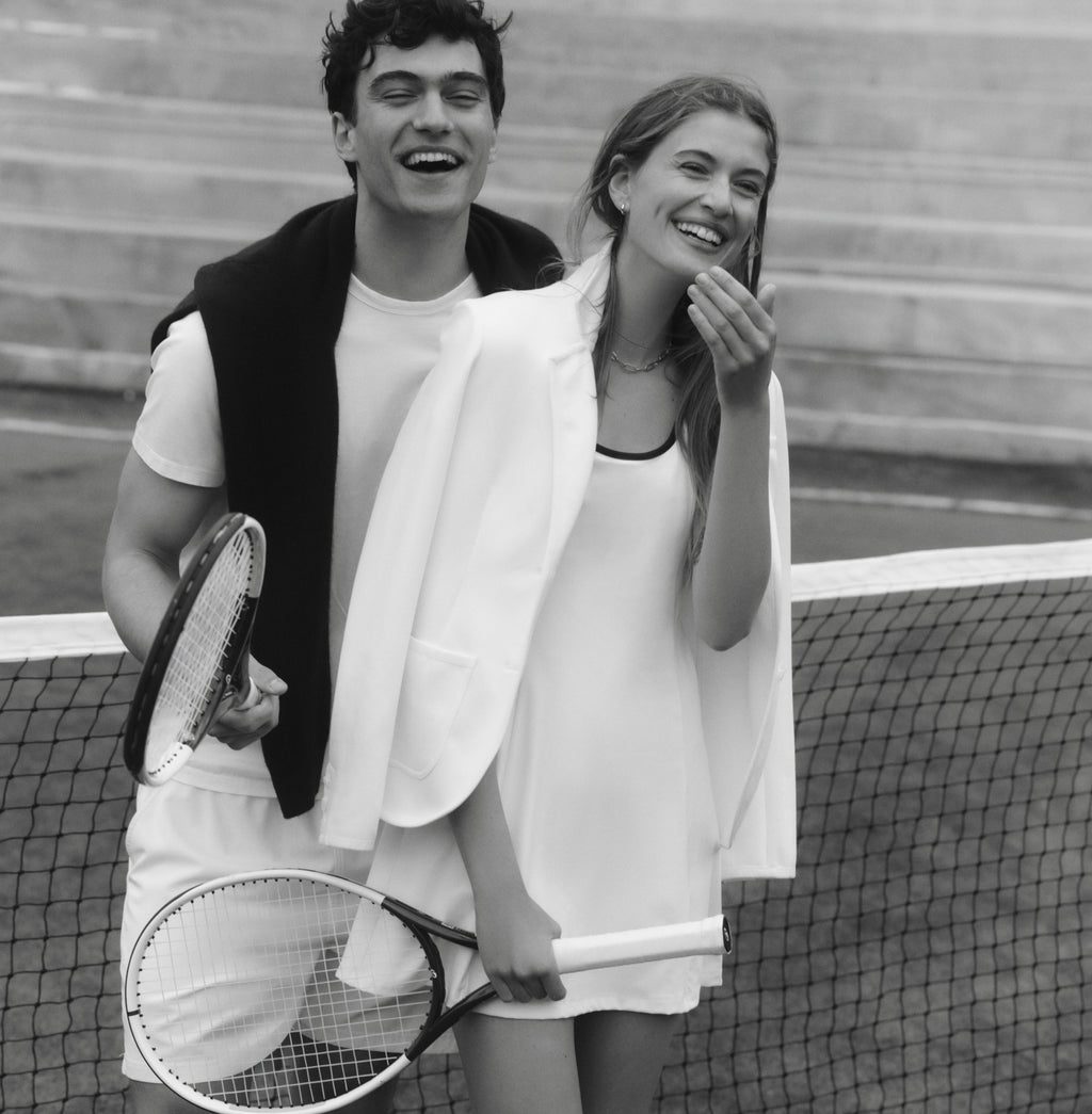 Black and white image of a couple on a tennis court. Man in a white tee and shorts, with a dark sweater over his shoulders. Woman in a white tennis dress and a white blazer.