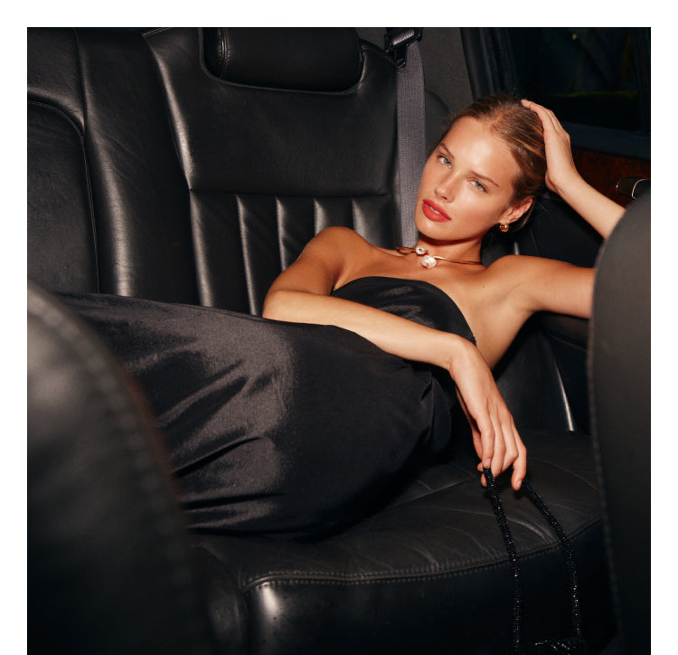 A woman wearing a black strapless maxi dress is sitting in a car with black leather seats.