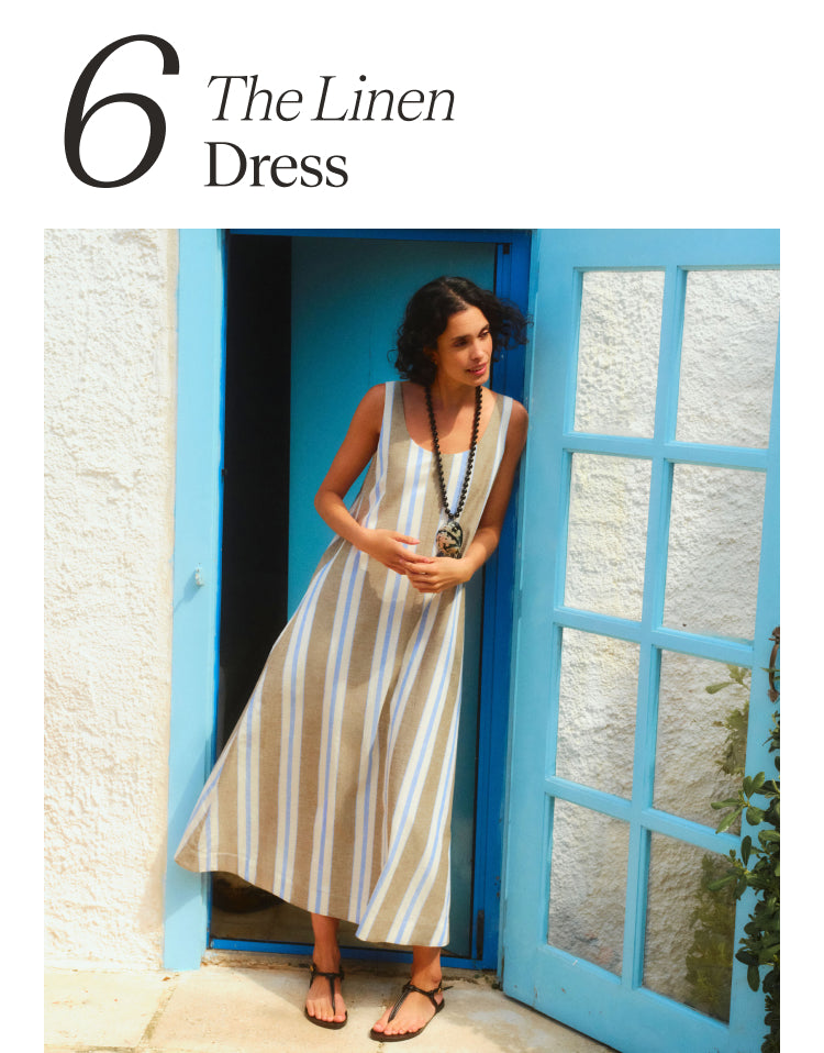 6 The Linen Dress. Woman standing inside a doorway in a long sleeveless dress with brown, blue, and white stripes.