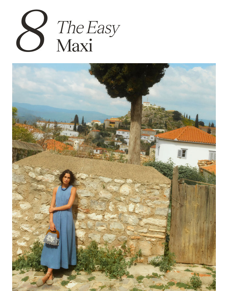8 the Easy Maxi Woman standing in a Greek village with terracotta topped homes in the background. Woman is wearing a sleeveless blue dress and holding an embroidered bag.