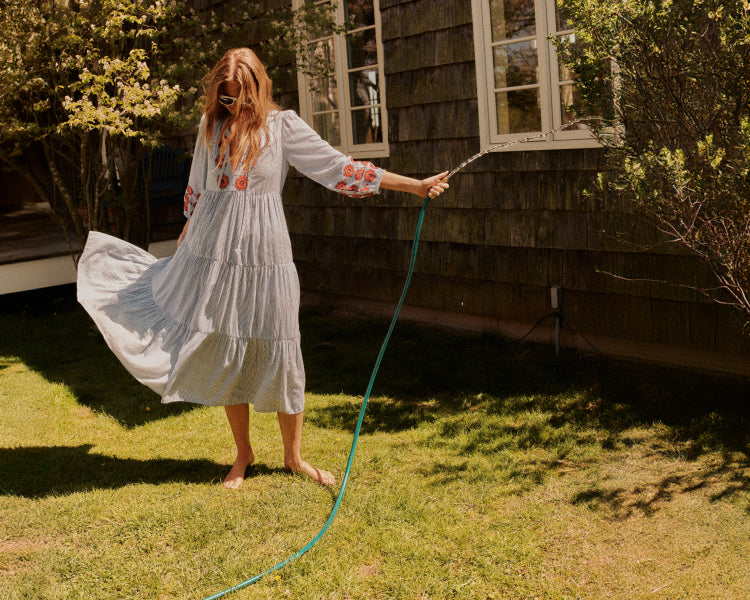 Woman in a long blue and white striped caftan with red flower details on the sleeves holding a hose.