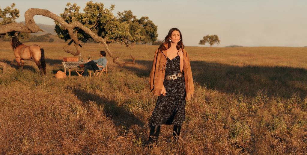 Woman in a field wearing a mid length slip dress with a belt , black leather boots, and a tan suede jacket with fringe. There's a man at a table, horse, and tree in the background.