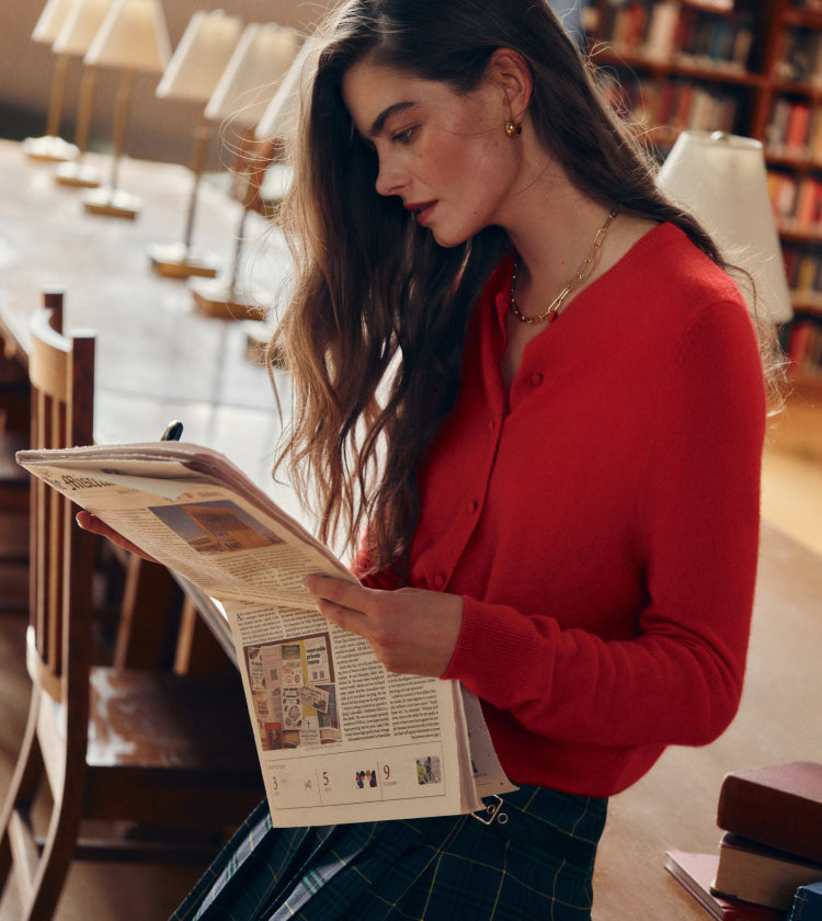 Woman reading a newspaper in a library in a red sweater and a blue plaid skirt.
