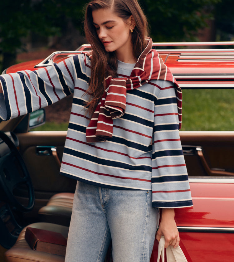 Woman opening a car door in a blue striped tee and jeans with a red striped tee draped over her shoulders.
