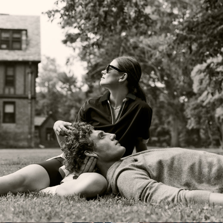 Man and woman sitting on grass. Woman is in a polo shirt with piping and sunglasses. Man is in a heathered sweater.