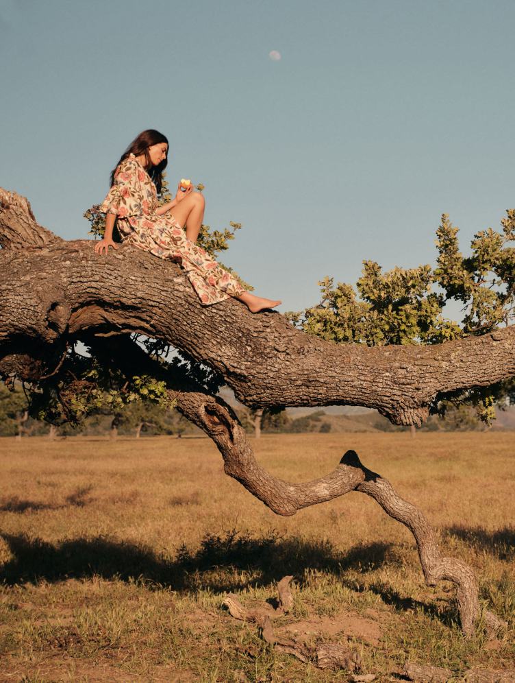 Woman lounging on a tree branch in a long floral dress with three quarter inch sleeves.