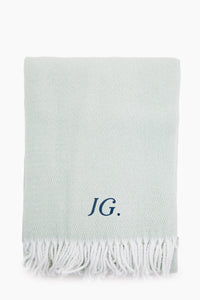 A light gray Matouk Opal Pezzo Throw features a white fringe and the initials JG. embroidered in dark blue on the front. Made from soft cotton, this elegant blanket adds style to any space.