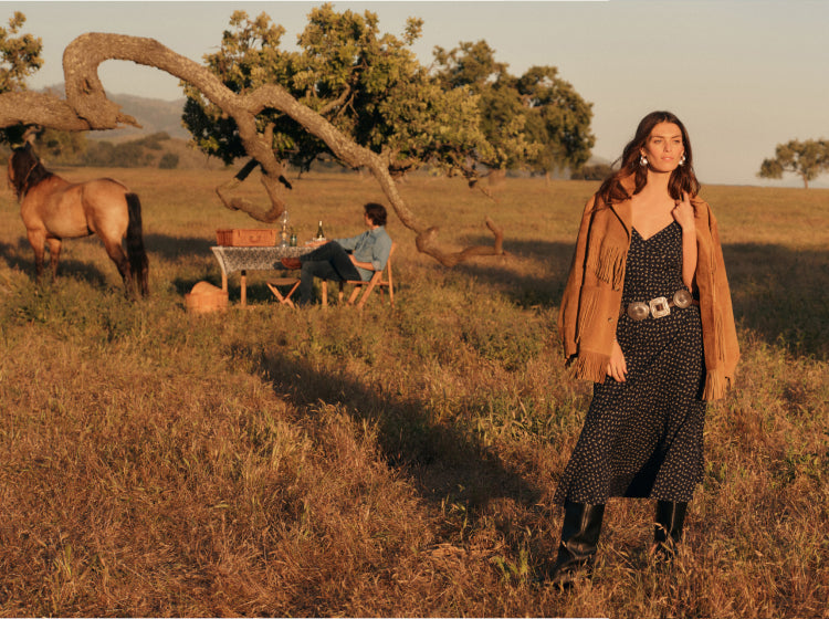 Woman in a field wearing a mid length slip dress with a belt , black leather boots, and a tan suede jacket with fringe. There's a man at a table, horse, and tree in the background.