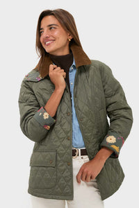 A woman smiles in a Barbour x Tuckernuck olive green quilted jacket with floral embroidered cuffs, paired with a blue shirt and white pants, standing against a plain background.