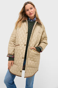 A woman wears the Barbour x Tuckernuck Miley quilt jacket, a beige diamond quilt style with brown trim, over a green sweater and blue jeans, standing with one hand in her pocket against a plain background.