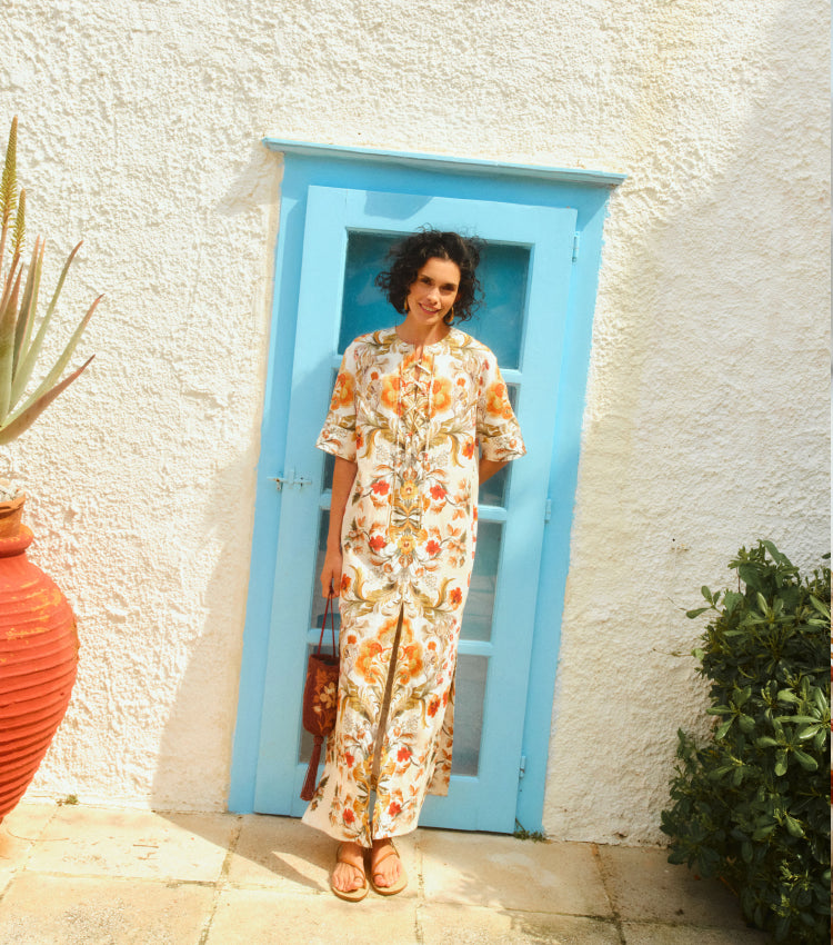 Woman standing in front of a blue door in a long, floral printed caftan with tan leather sandals.
