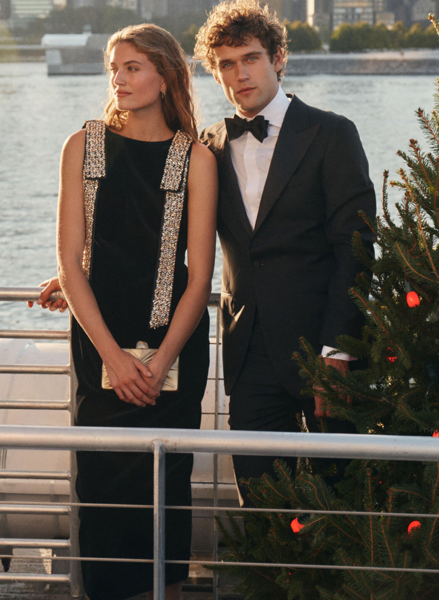On the left, a woman wears a black midi dress with embellished shoulder bows and carries a gold clutch. On the right, a man wears a suit and bow tie next to a Christmas tree.