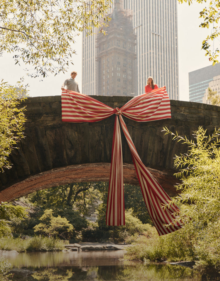 A man and a woman standing on a city park bridge holding an oversized red and white striped bow.