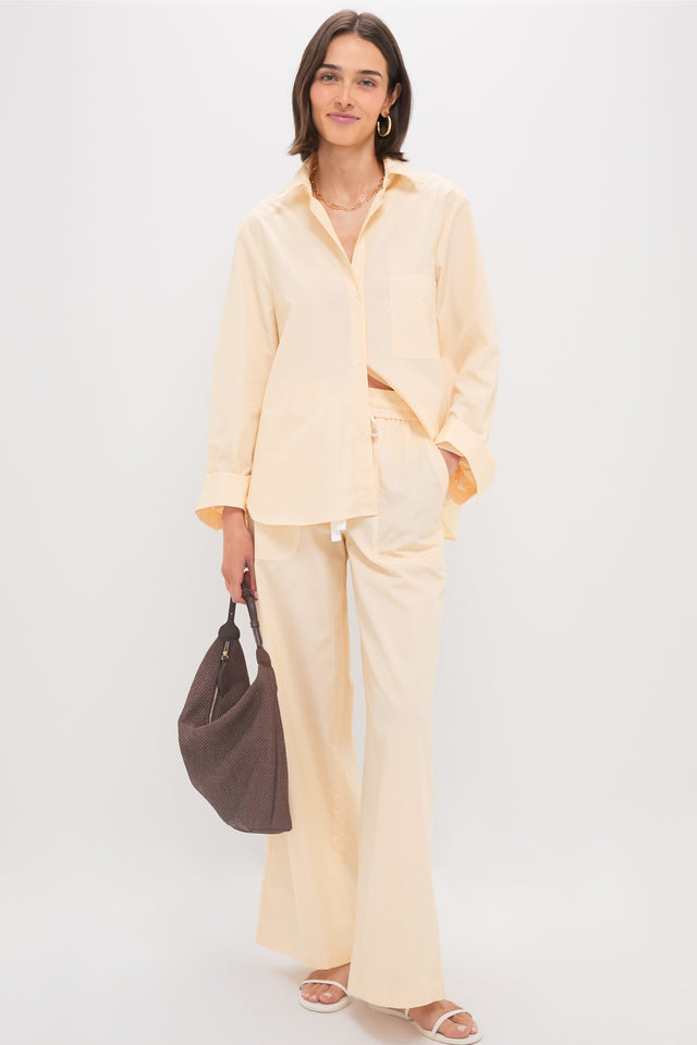 A person stands against a plain background wearing the TWP Next Morning Shirt in Ivory and Yellow with an oversized fit, paired with matching wide-leg pants. The look is completed with a dark brown bag, white sandals, and is made from 100% cotton.