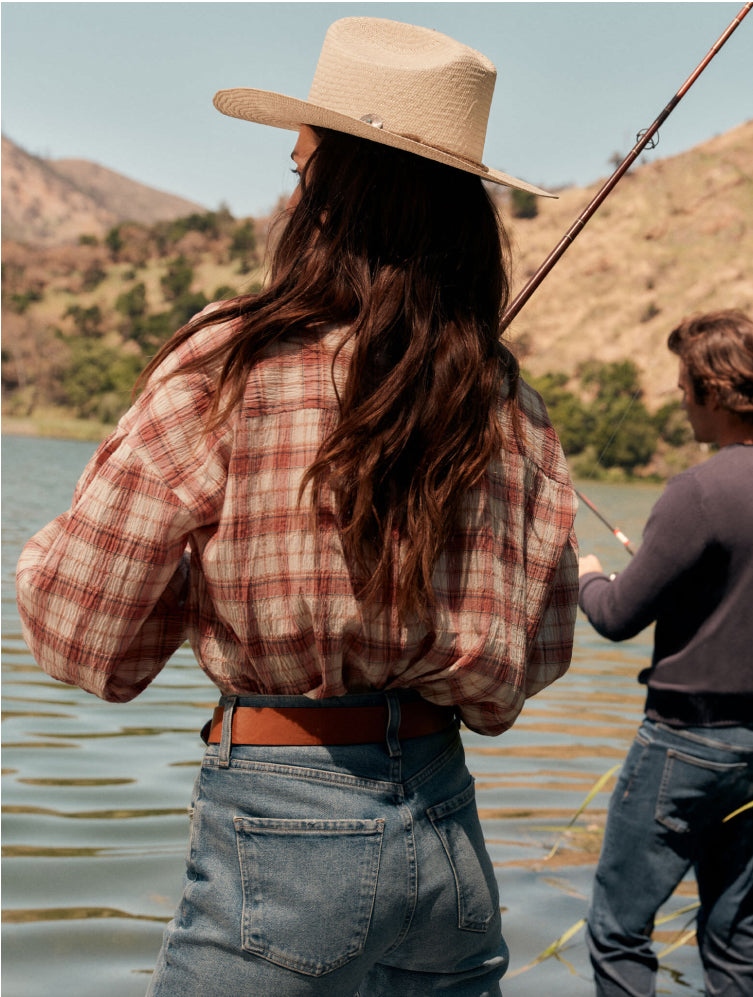 The back of a woman fishing in a straw hat, plaid shirt, and jeans.
