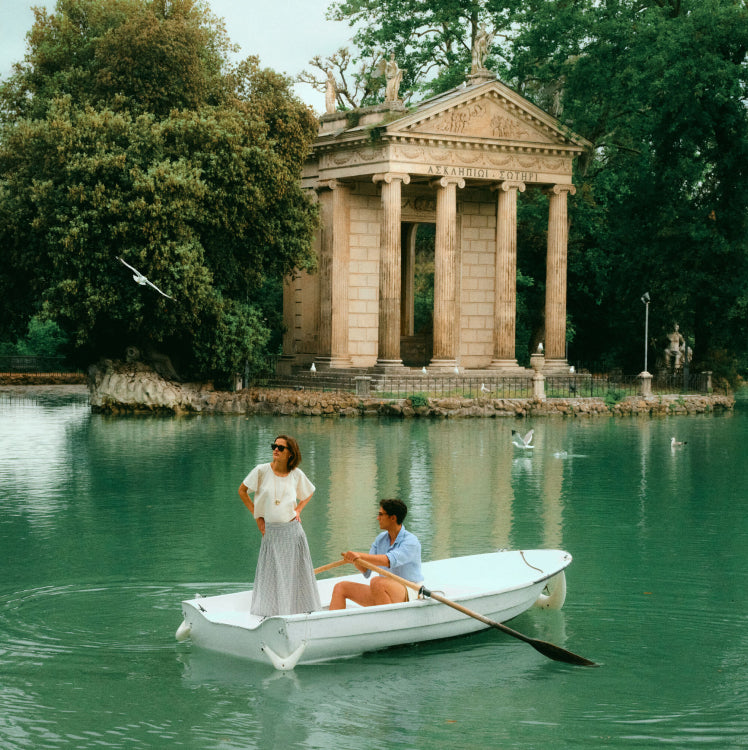 Man and woman in a boat in the middle of the water near an archaic building. Woman in a white top and a blue skirt.