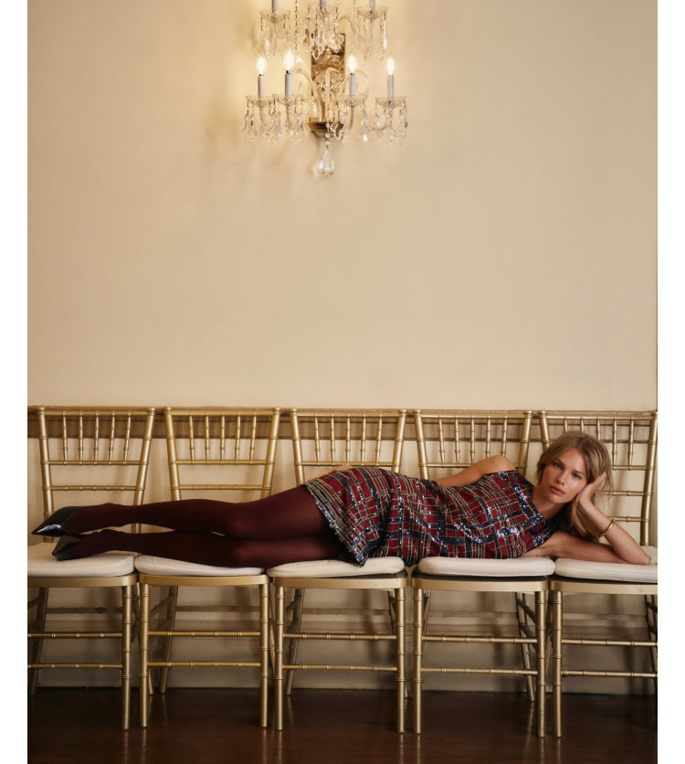 A woman wearing a plaid, sequin mini dress, black tights, and black pointed heels while lying across 5 chairs.