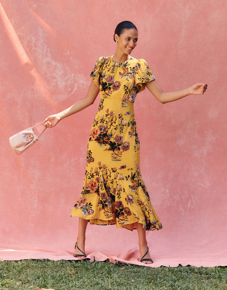 Woman in yellow floral midi dress standing in front of a pink background.
