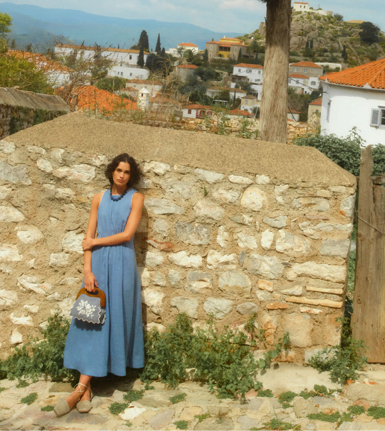 Woman standing in a Greek village with terracotta topped homes in the background. Woman is wearing a sleeveless blue dress and holding an embroidered bag.