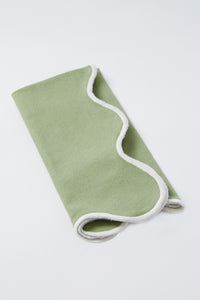 A folded light green Tuckernuck Home blanket with white trim, displayed on a white surface, pairs beautifully with scalloped napkins for an elegant touch.