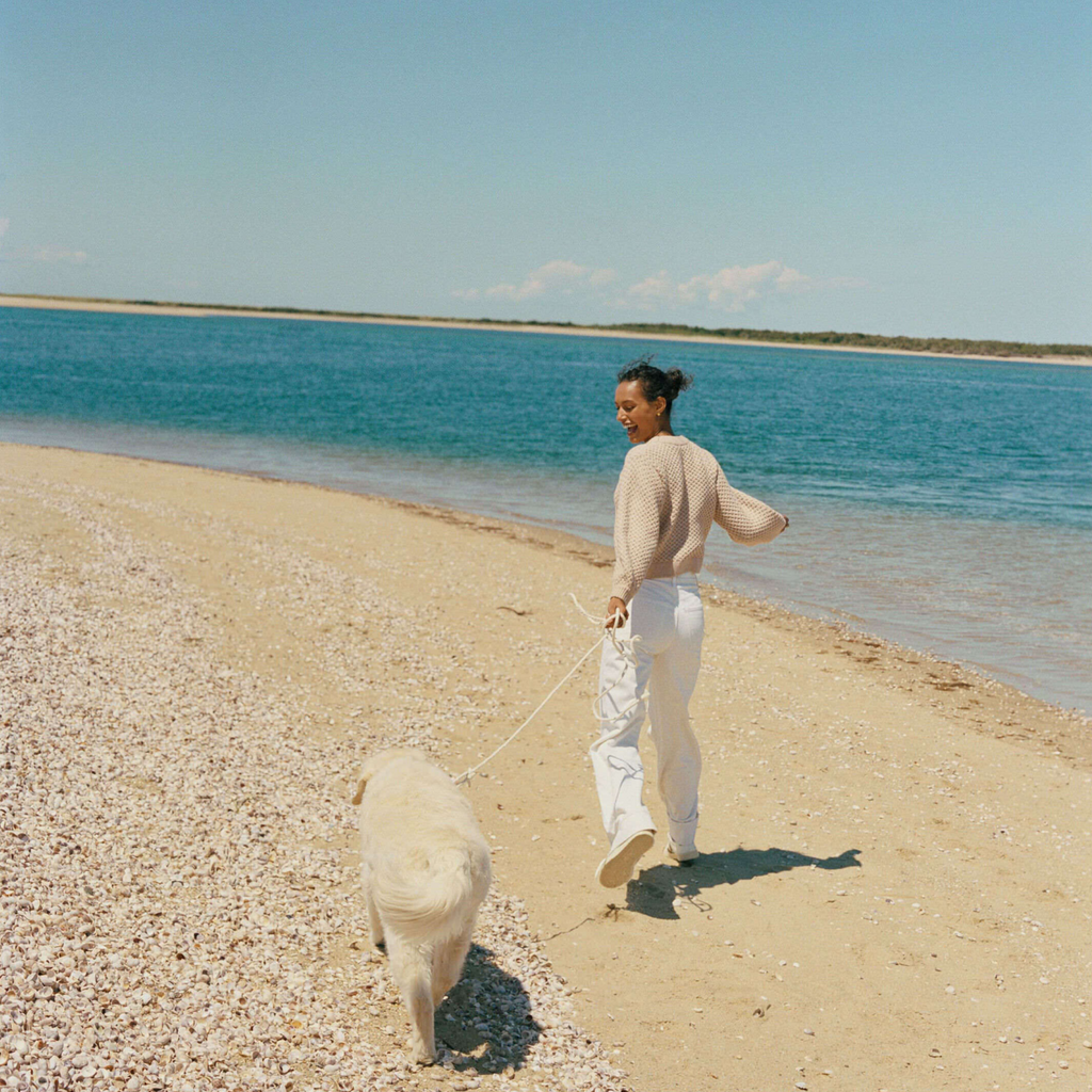 Person in light trousers and knit sweater walking a light-colored dog on a shell-strewn beach, embodying classic, effortless, fun style at the water's edge.