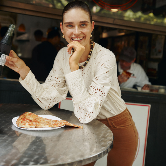A young person in a classic, embroidered blouse enjoys an effortless, fun moment with pizza and a soda, embodying Tuckernuck's high-end, relaxed style.