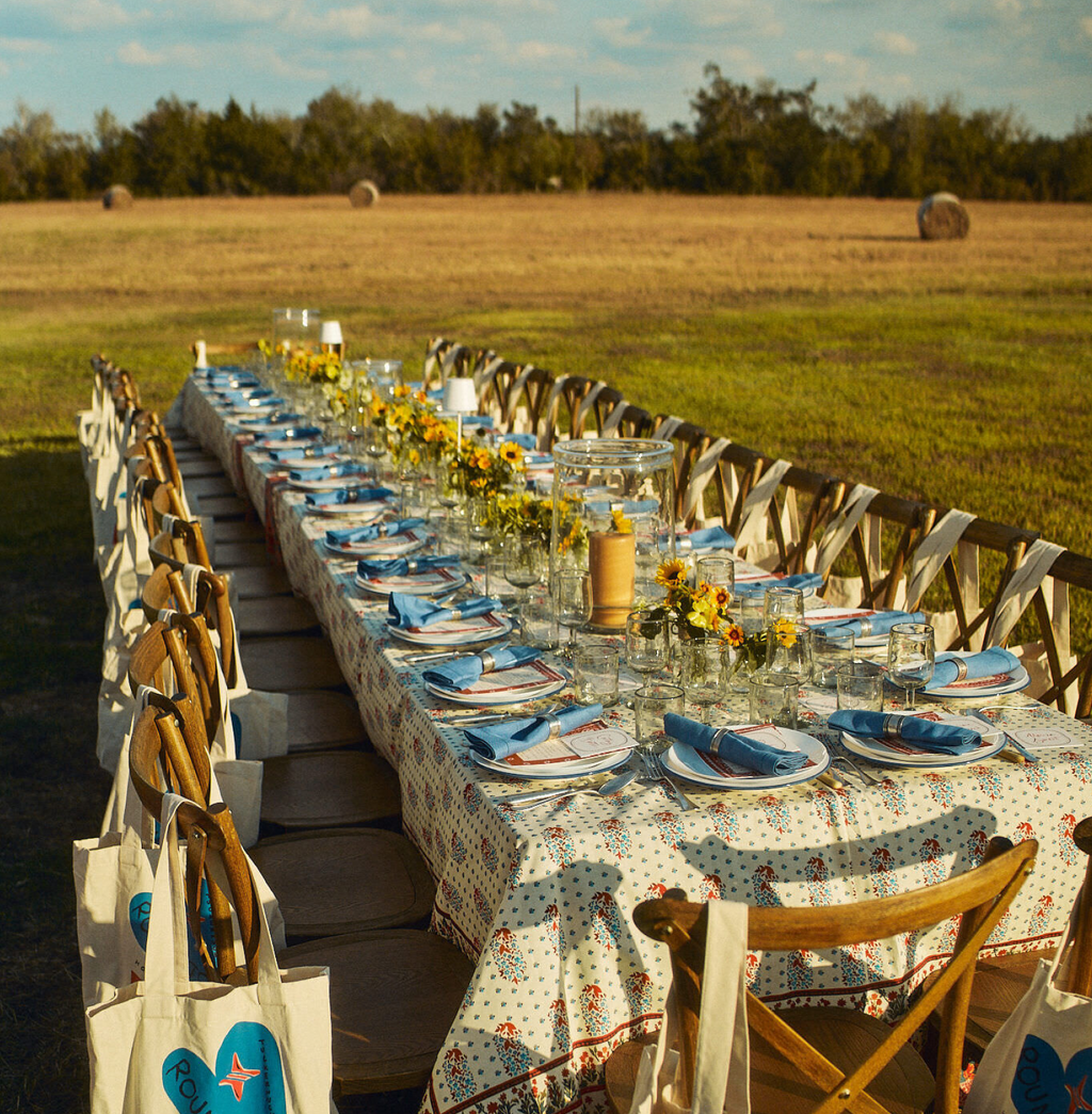 Outdoor dining scene with a classic farm-style table, patterned tablecloth, elegant place settings, and yellow floral arrangements, exuding effortless high-end charm and fun ambiance.
