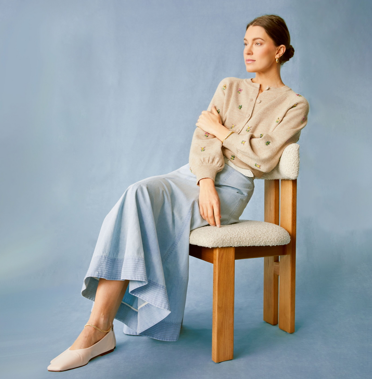 Young woman in a classic, embroidered beige cardigan, pale blue flared denim skirt, and nude flats, exuding effortless, high-end style with minimal gold jewelry.
