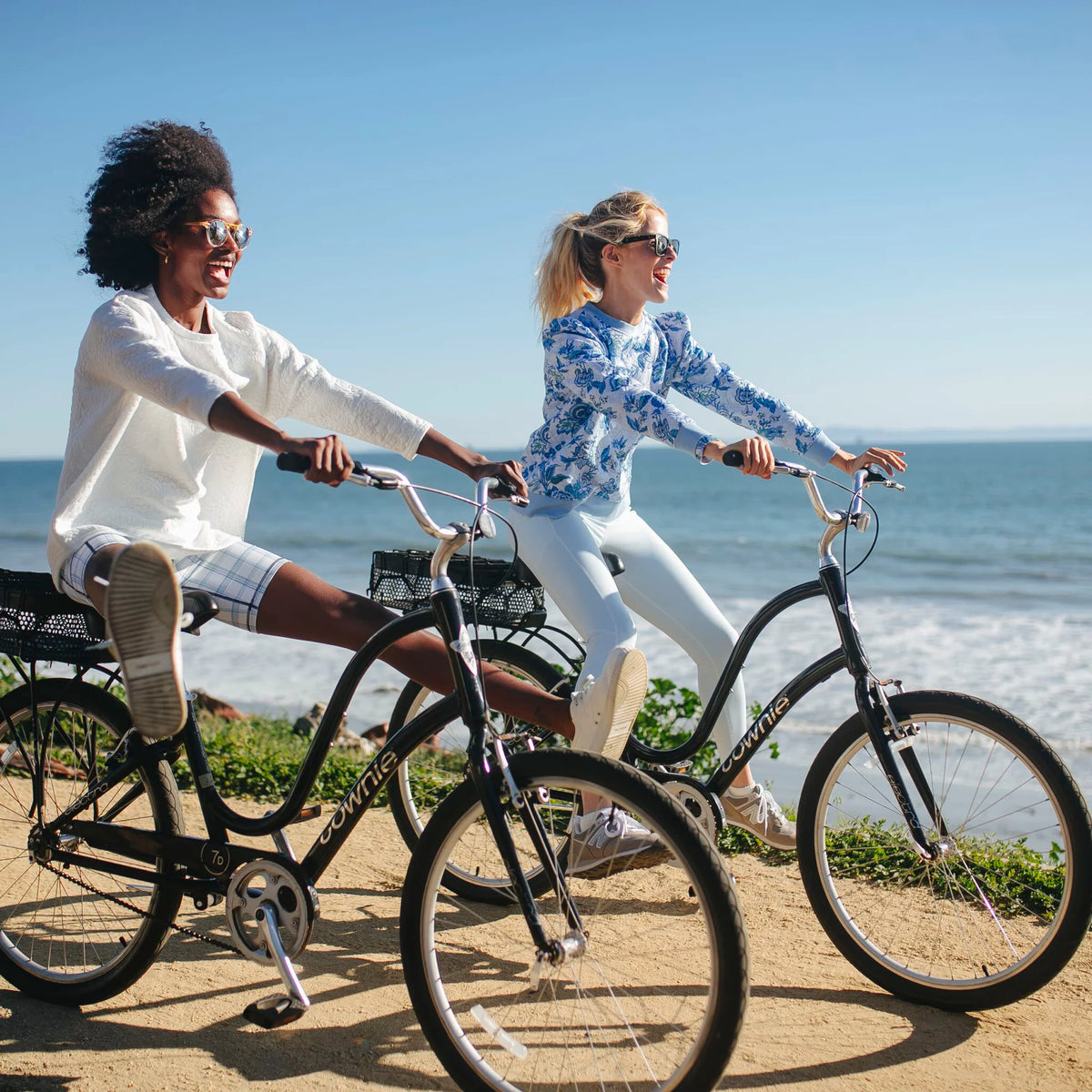 Two women joyfully ride black cruiser bicycles along the beach, showcasing effortless style with classic, fun outfits from Tuckernuck's high-end collection.