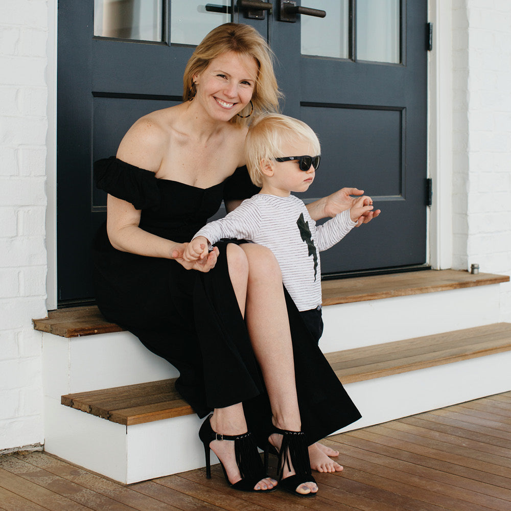 Woman in a classic black off-the-shoulder dress and tassel sandals sits with a toddler in fun striped attire, embodying effortless, high-end style on a porch.