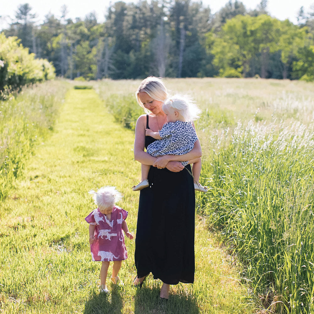 Woman in a classic black dress carries a child in a blue dress, walking through a sunlit meadow, embodying effortless, high-end, fun style.
