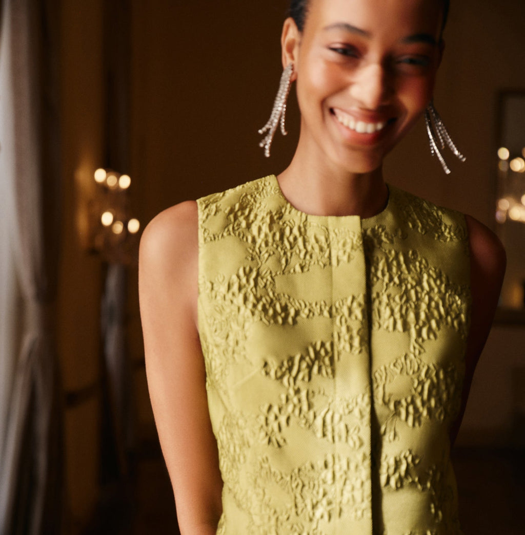 Close-cropped portrait of a smiling woman in a classic, pale green textured dress, with sparkling drop earrings; exudes an effortless, fun, high-end elegance.